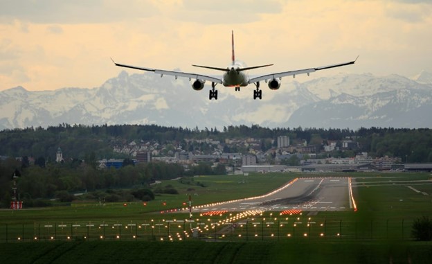 Flyg som ska landa på en upplyst landningsbana. Framför syns ett samhälle och snötäckta berg.