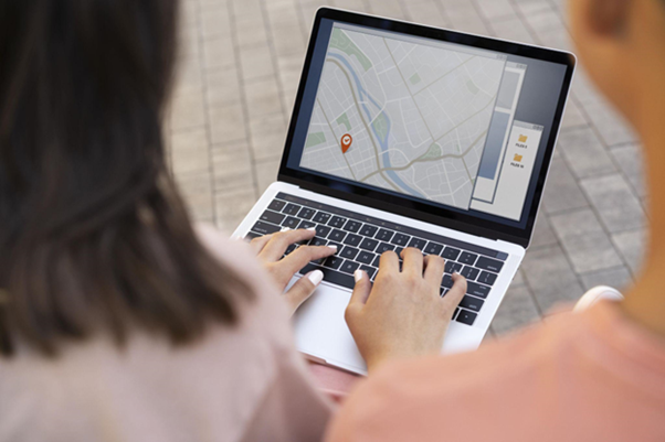 Two people sitting with their backs to the camera, using a laptop to track a shipment. In the background, stone pavement is visible.