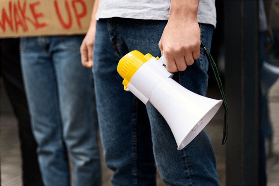 A person is holding a megaphone down by their leg. In the background, you can see another person's legs and a sign that says "Wake up".