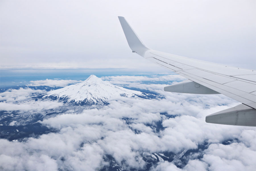 A view from an airplane showing the wing of the aircraft in the foreground, surrounded by white clouds enveloping a snow-covered mountain in the background.