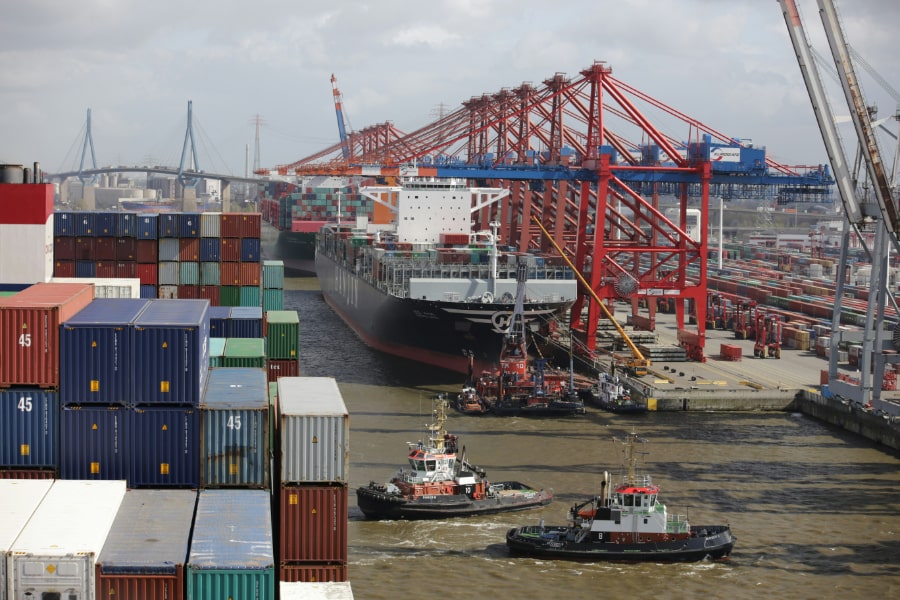 Containerfartyg med färgglada containrar som lotsas till kajplats. Fartyg lastas med kranar i bakgrunden.