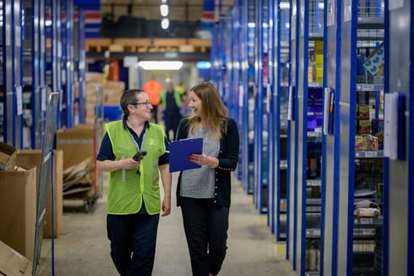 Two women walking side by side along tall blue shelves in a warehouse. They are smiling at each other.