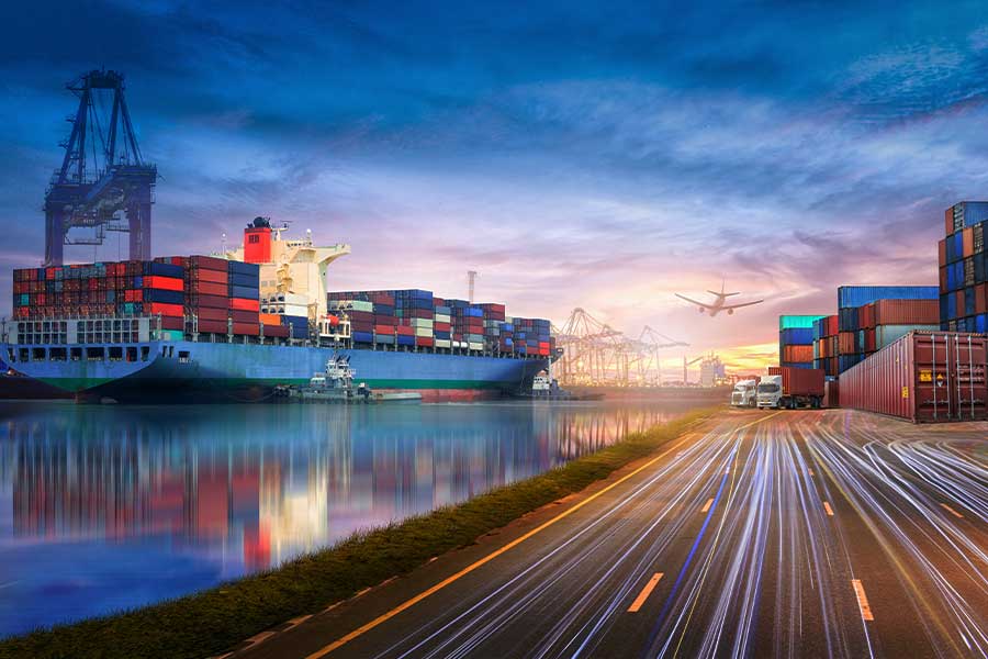 An airplane flying over a container port and a container ship with crains in the background